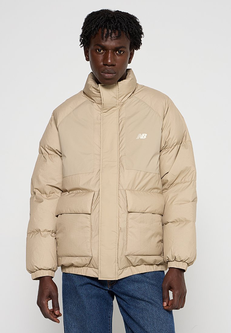 Chaqueta acolchada beige con cuello alto, bolsillos frontales y un logo simple en el pecho. Está hecha de un material ligero con una textura suave.