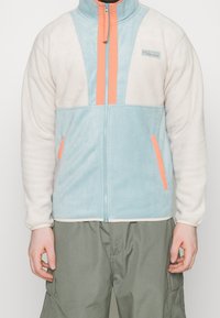 Giacca in pile di colore azzurro chiaro e crema con strisce verticali arancioni, con chiusura a zip completa e tasche laterali. Collo arrotondato e texture morbida.