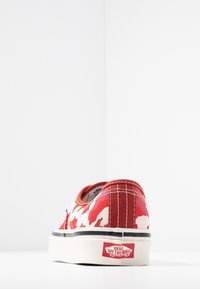 Baskets en toile rouge et blanche avec un motif camouflage, semelle en caoutchouc noire, et coutures blanches. Logo Vans visible sur le talon.