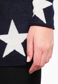 Hand mit blassem Nagellack neben einem nav blauen Pulloverärmel, der ein großes weißes Sternmuster auf einem weißen Hintergrund zeigt.