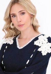 Marineblauer Pullover mit V-Ausschnitt, verziert mit weißen gehäkelten Blumenakzenten an den Schultern und flauschigem weißen Saum entlang des Ausschnitts und des Saums.