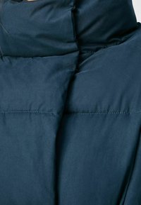 Marinblå pufferjacka med hög krage, slät textur och horisontella quiltade sektioner. Synliga sömnadsdetaljer längs sömmarna.