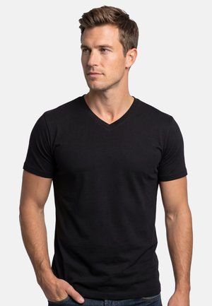 Junger Mann mit kurzen braunen Haaren, der ein schwarzes V-Ausschnitt-T-Shirt trägt, mit einer Hand in der Tasche stehend und nach links schauend.