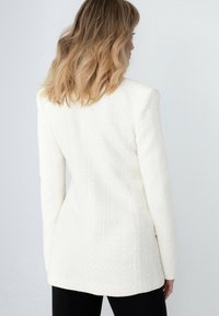 Bílý tvídový blazer s texturovaným povrchem, prodlouženou siluetou a strukturovanými rameny, stylizovaný s černými kalhotami.