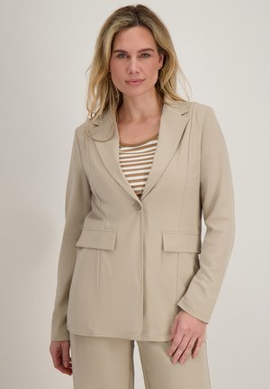Blonde vrouw met beige op maat gemaakte blazer met klepzakken en beige gestreept topje, staand voor een effen achtergrond.