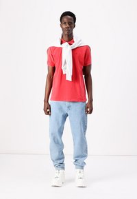 Polo rouge à manches courtes avec un pull blanc drapé sur les épaules, associé à un jean baggy bleu clair et des sneakers blanches.