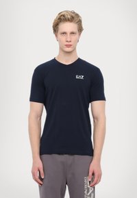 Námořnická modrá V-neck trička vyrobená z měkké látky, s malým bílým logem EA7 na hrudi. Model má na sobě šedé tepláky s logem.