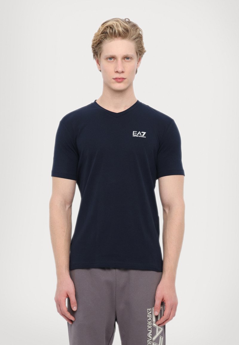 Námořnická modrá V-neck trička vyrobená z měkké látky, s malým bílým logem EA7 na hrudi. Model má na sobě šedé tepláky s logem.