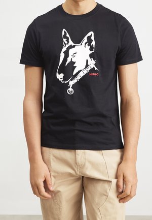 Homme portant un t-shirt noir avec un motif tête de chien blanc et texte rouge "HUGO,", associé à un pantalon beige.