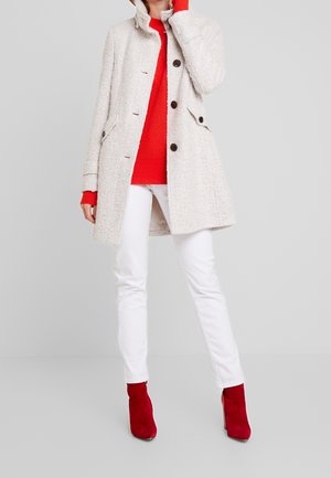 Femme portant un manteau clair texturé, un pull rouge, un pantalon blanc et des bottines rouges, debout devant un fond gris uni.