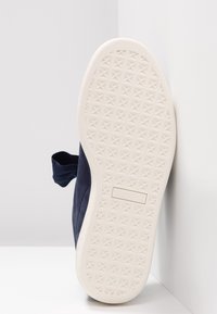 Semelle en caoutchouc blanche texturée d'une chaussure bleu foncé reposant contre un mur blanc, avec une languette à l'arrière.