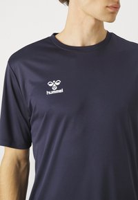 Tummansininen urheilupaita, joka on valmistettu sileästä kankaasta, ja siinä on pyöreä kaula-aukko sekä valkoinen "hummel"-logo vasemmassa rintakohdassa.