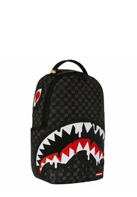 Mochila de cuadros negros con un gran diseño de boca de tiburón en blanco, rojo y negro en la parte frontal, y un parche rojo y blanco en el costado.