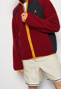 Rote Fleecejacke mit schwarzen Stoffakzenten, gelbem Reißverschluss und Logo. Das Design verfügt über einen hohen Kragen und lange Ärmel für zusätzliche Wärme.