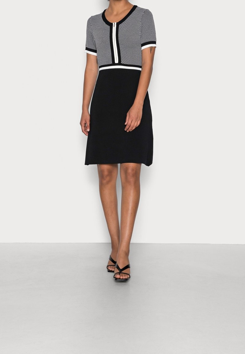 Femme portant une robe courte à manches courtes texturée noire et blanche avec une fermeture éclair à l'avant et des talons noirs à lanières, debout devant un fond uni.