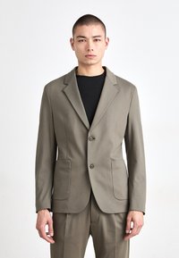 Beige blazer med en skreddersydd passform, notklapper, to knapper og påsydd lommer. Båret over en svart t-skjorte laget av glatt stoff.