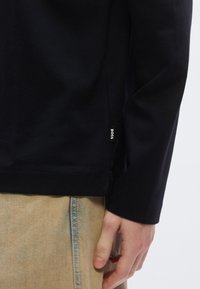 Črna majica z dolgimi rokavi iz bombaža, z stranskim razporkom in subtilno etiketo z napisom »BOSS«, kombinirana s svetlim denimo.