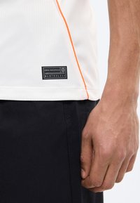 Κοντινό πλάνο ενός χεριού δίπλα στο τελείωμα ενός λευκού πουκαμίσου με πορτοκαλί ραφή και μαύρη ετικέτα "Engineered" με σειριακούς αριθμούς, φοράει μαύρο παντελόνι.