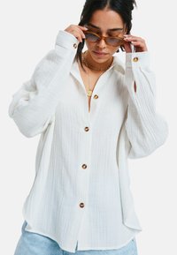 Witte button-up shirt met textuurstof, voorzien van grote bruine knopen en lange mouwen met opgerolde manchet. Draagt het met lichtblauwe jeans.