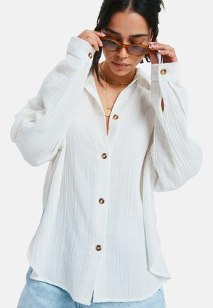 White button-up shirt with textured fabric, featuring large brown buttons and long sleeves with rolled cuffs. Worn with light blue jeans.