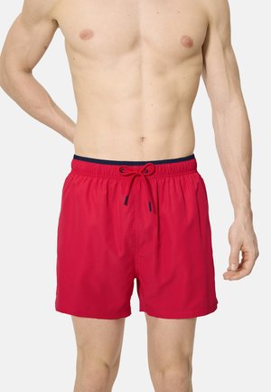Torso masculino vistiendo pantalones cortos de baño rojos con cintura azul marino y cordón rojo, de pie contra un fondo claro y liso.
