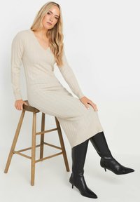 Beige geripptes Strickkleid mit V-Ausschnitt und langen Ärmeln, kombiniert mit knielangen schwarzen Lederstiefeln mit spitzem Zehenbereich. Modell sitzt auf einem Holzstuhl.