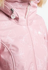 Veste rose clair avec motifs floraux blancs, dotée d'un col montant, d'une fermeture éclair frontale et d'un petit logo près de l'ourlet. Texture douce.