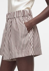 Άτομο που φοράει σορτς με ψηλή μέση και κάθετες καφέ και λευκές ρίγες, χέρι στην πλαϊνή τσέπη, σε συνδυασμό με ανοιχτόχρωμο τοπ.