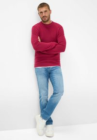 Maglione rosso a maglia con scollo rotondo, abbinato a jeans azzurri chiari e sneaker bianche, in piedi su uno sfondo bianco uniforme.