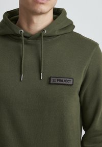 Olive green cotton hoodie with a front kangaroo pocket, adjustable drawstrings, and a textured black patch reading "11 PROJECT."