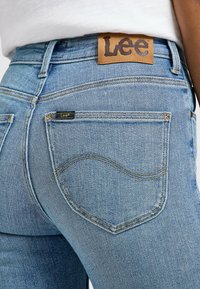Jeans de mezclilla azul claro con una etiqueta de cuero que presenta "Lee" en la cinturilla. Costuras distintivas y dos bolsillos traseros visibles.