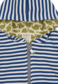Felpa a righe blu e bianche con zip grigia. All'interno presenta una fodera verde con motivi di balene bianche. Realizzata in cotone 100% biologico.