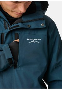 Türkisfarbene wasserdichte Jacke mit durchgehendem Reißverschluss, verstellbaren Bündchen und einer Brusttasche. Mit gesticktem Logo und verstärkten Nähten.