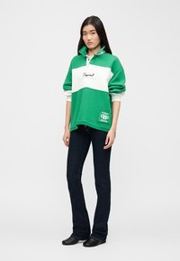 Νεαρή γυναίκα όρθια, φορώντας μακρυμάνικο πουλόβερ σε πράσινο και λευκό με λογότυπο, σκούρο μπλε τζιν και μαύρα παπούτσια, μπροστά σε απλό ανοιχτόχρωμο φόντο.