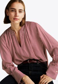 Rød og hvid mønstret bluse med v-halsudskæring, plisserede detaljer og lange ærmer. Matchet med mørke jeans og et mørkt bælte.