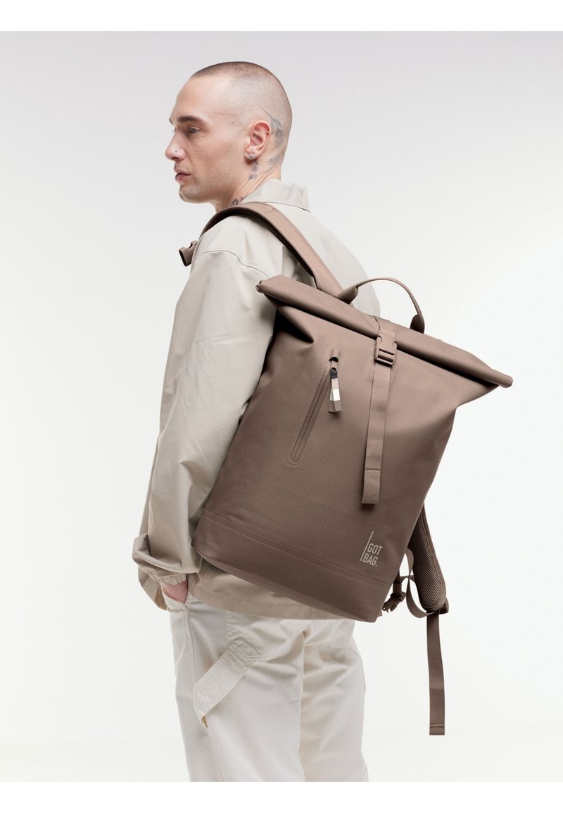 Sac à dos roll-top marron en matériau durable, avec des straps réglables, une poche avant et un logo sur le côté.