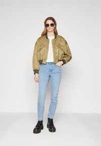 Jachetă bomber verde măsliniu cu fermoar frontal, două buzunare aplicate, mansete ribificate, combinată cu blugi skinny de culoare albastru deschis și cizme negre cu șireturi.