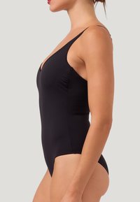 Kvinna iklädd en svart baddräkt med tunna axelremmar, visad i profil mot en enkel ljus bakgrund.