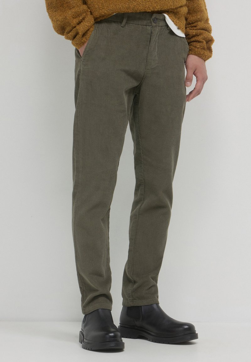 Olivgrüne Cordhosen mit schmaler Passform. Verfügt über Seitentaschen und einen Knopfverschluss, kombiniert mit schwarzen Knöchelstiefeln.