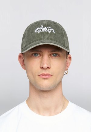 Uomo con occhi azzurri che indossa un berretto verde oliva con testo bianco ricamato, una maglietta bianca, un orecchino a cerchio in oro e una sottile collana d’oro.