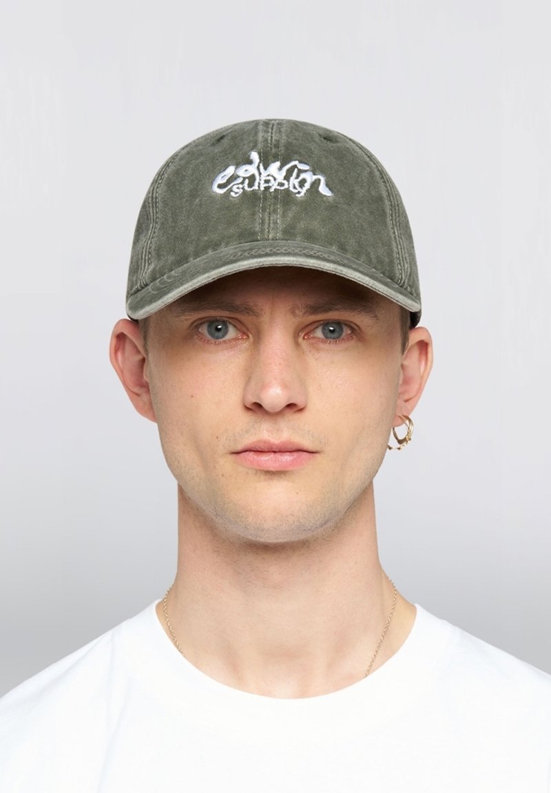 Hombre con ojos azules que lleva una gorra verde oliva con texto bordado en blanco, camiseta blanca, pendiente de aro dorado y collar delgado de oro.