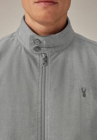 Hombre vestido con una chaqueta gris con cremallera y cuello alto, abrochada con dos botones y un pequeño logotipo de ciervo bordado en el pecho.