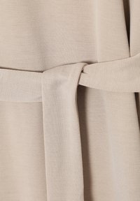 Profilo di tessuto beige con texture e dettaglio della cintura annodata. Il tessuto ha un motivo intrecciato sottile ed è leggero, creando una caduta morbida.