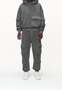 En person som bär en mörkgrå hoodie och matchande cargo-byxor med flera fickor, ihop med vita sneakers, stående mot en enkel bakgrund.