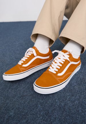 Osoba w beżowych spodniach i białych skarpetkach, nosząca niskie pomarańczowe trampki z białymi sznurowadłami i bocznym paskiem, siedzi na ciemnoniebieskim dywanie.