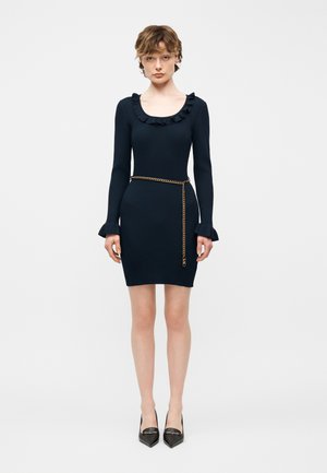 Femme portant une robe noire en maille côtelée avec un col et des poignets à volants, accessoirisée d'une ceinture chaîne en or et de talons noirs à bout pointu.