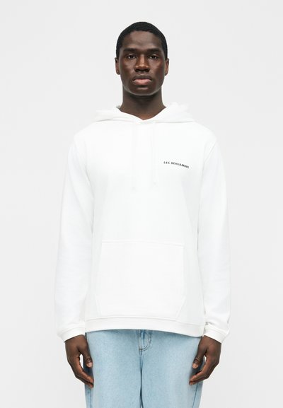 Hombre de pie mirando al frente, vestido con una sudadera blanca con el texto "LES BENJAMINS" y vaqueros azul claro sobre un fondo liso.