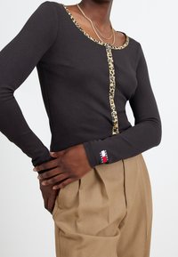 Svart långärmad topp med leopardmönstrad halsringning och knappparti, i kombination med beige byxor; tyget verkar slätt och figurnära.