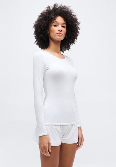 Γυναίκα με σγουρά μαλλιά, που φορά στενό λευκό μακρυμάνικο τοπ και λευκά σορτς, στέκεται μπροστά σε απλό φωτεινό φόντο.