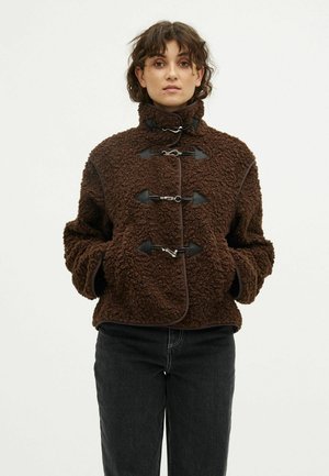 Woman wearing a brown textured fleece jacket with toggle closures and black pants, standing with hands in pockets against a plain background.